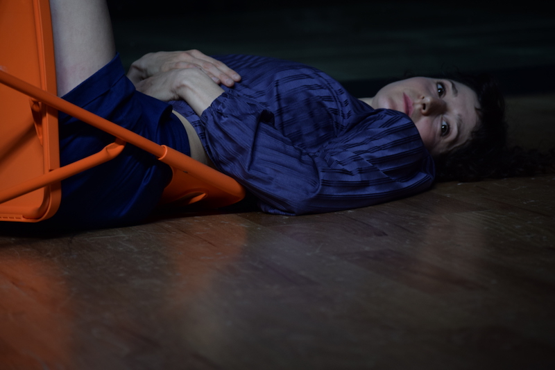 Gabrielle lays in a orange folding chair on the ground. She gazes toward the camera.